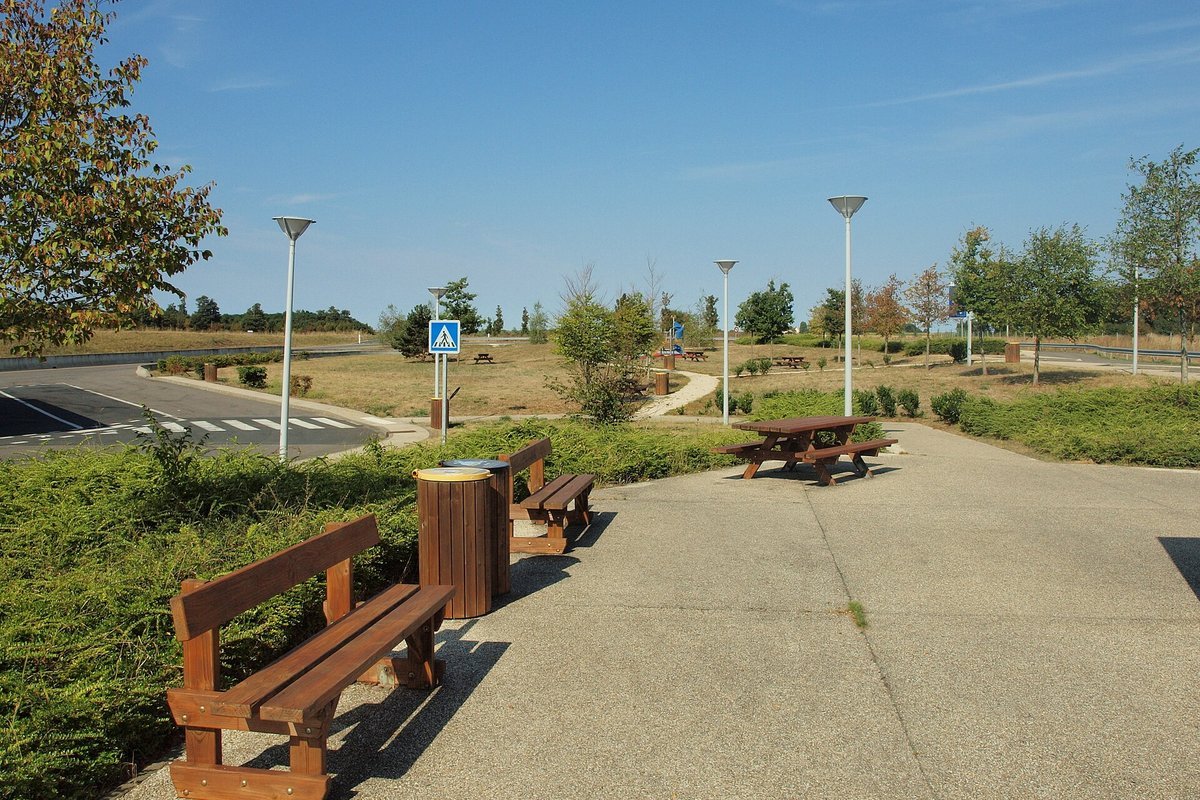 Aire de repos d’autoroute avec bancs en bois, poubelle et tables de pique-nique au bord d’une allée, sous un ciel bleu