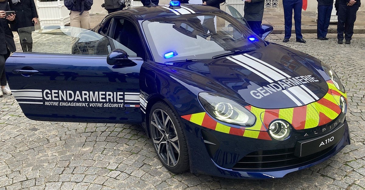 Alpine A110 bleu nuit aux couleurs de la gendarmerie nationale, porte ouverte, avec bandes jaunes et rouges à l’avant et gyrophares bleus allumés, garée sur des pavés