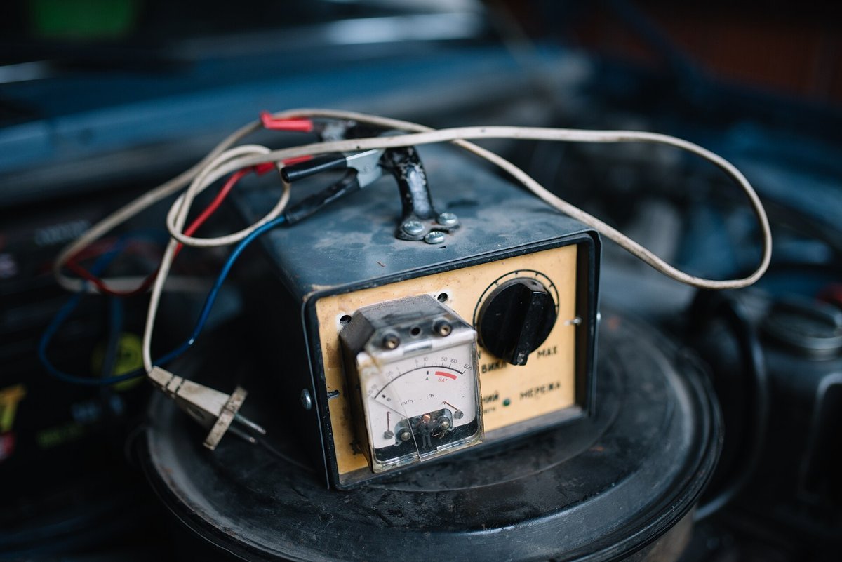 Ancien chargeur de batterie à cadran et gros bouton, posé sur un bidon noir dans un garage, câbles et pinces autour
