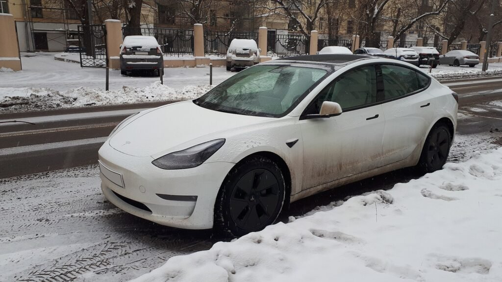 Voiture électrique blanche aux jantes noires garée le long d’un trottoir enneigé dans une rue de ville, en plein hiver