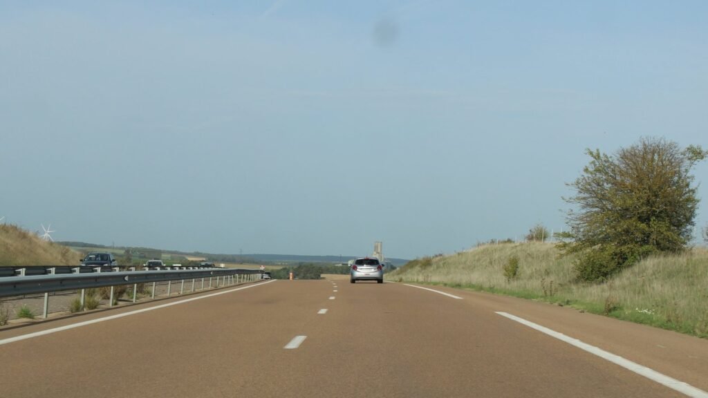 Voies de l’autoroute A6 en octobre : une voiture grise roule sur la voie de droite sous un ciel bleu, avec des éoliennes au loin