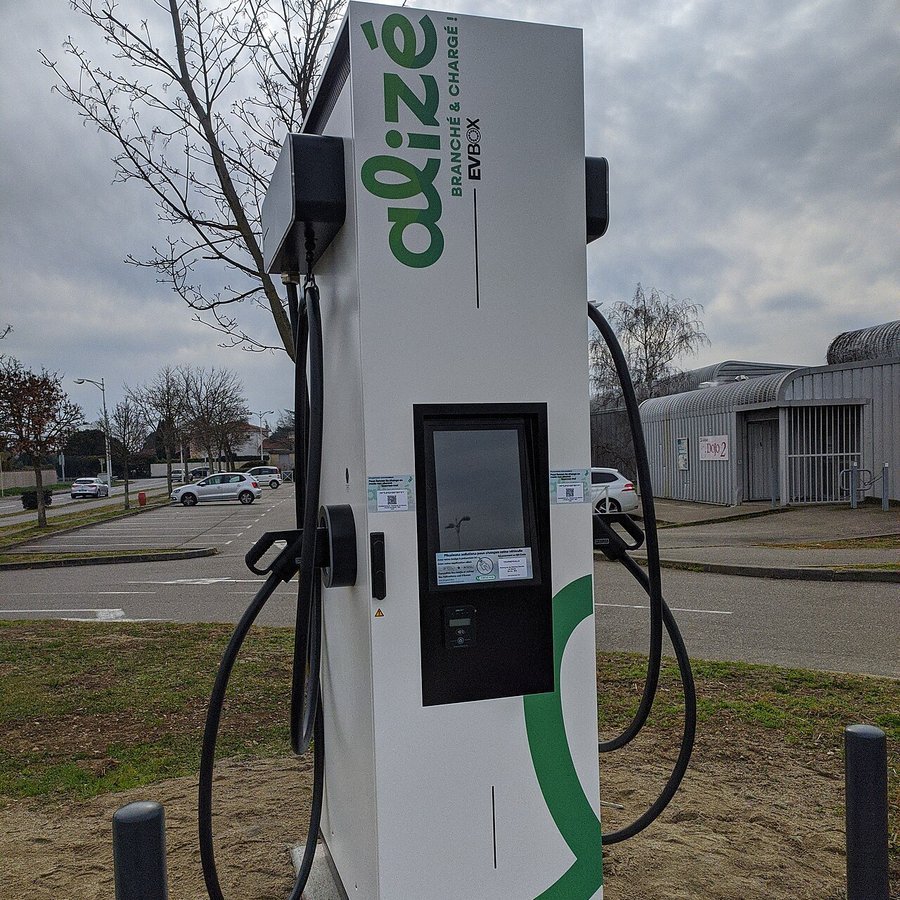 Borne de recharge rapide blanche et verte du réseau Alizé, avec écran et câbles, sur un parking de Tournefeuille