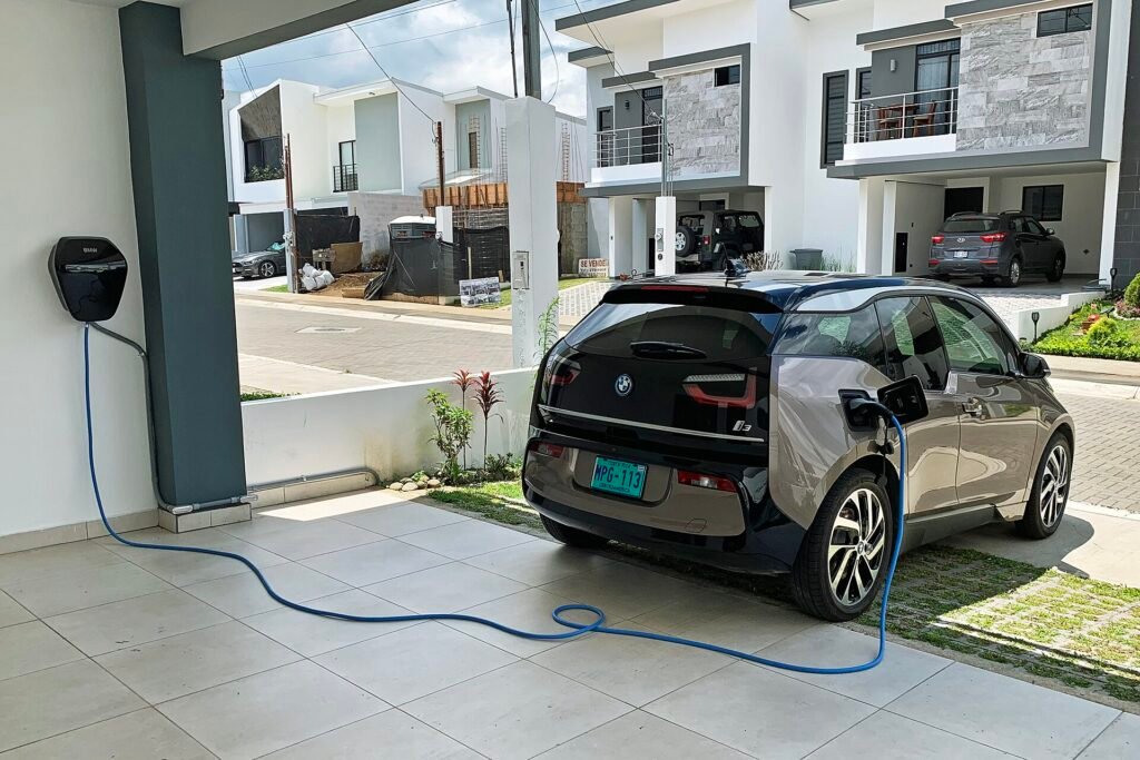 Voiture électrique grise branchée par un câble bleu à une borne de recharge murale fixée sous l’abri d’une maison