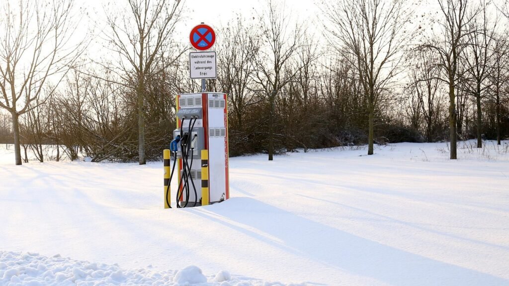 Borne de recharge rapide pour voitures électriques au milieu d’un parking couvert de neige, sous un soleil d’hiver, devant des arbres sans feuilles