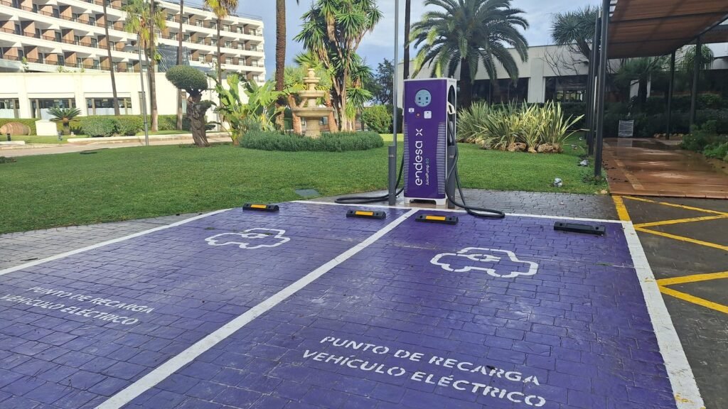 Borne de recharge Endesa X entre deux places violettes marquées « Punto de recarga vehículo eléctrico », sur un parking bordé de pelouse et de palmiers, devant des hôtels à Cala Millor (Majorque)