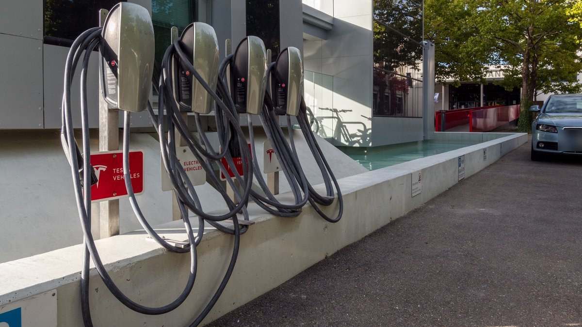Quatre bornes murales Tesla avec leurs câbles enroulés devant un hôtel, panneaux rouges indiquant des places réservées aux Tesla et une place ouverte aux autres véhicules électriques