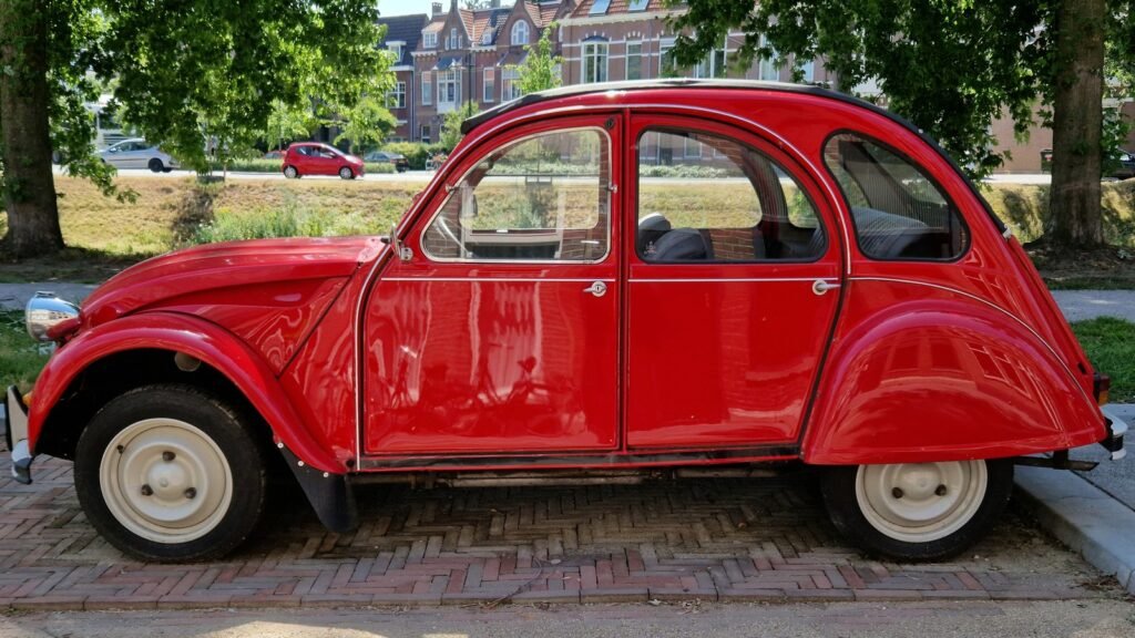 Une Citroën 2 CV rouge d’époque, vue de profil, garée sur un pavage de briques devant des arbres et des maisons en brique