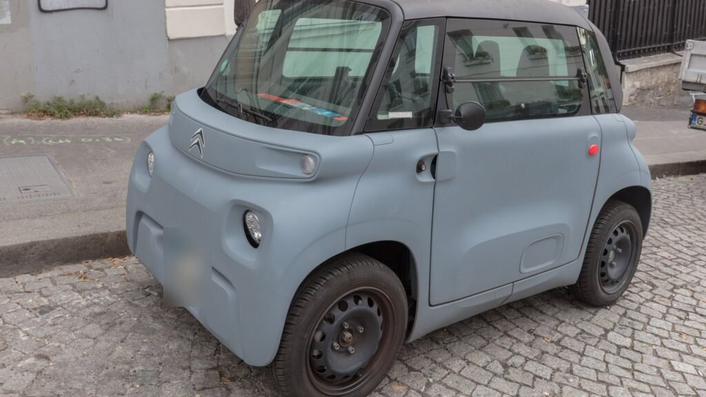 Citroën Ami gris bleu, petite voiture sans permis électrique, garée sur des pavés dans une rue de Paris, vue de trois quarts avant