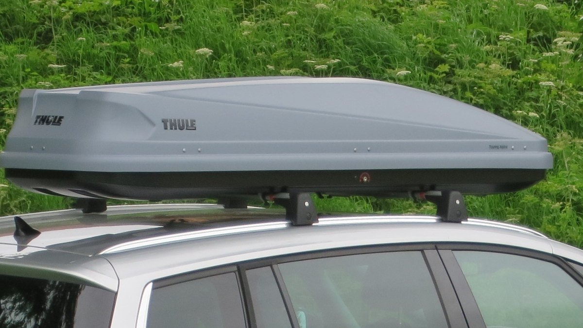 Coffre de toit Thule gris fixé sur deux barres transversales, elles-mêmes montées sur les barres longitudinales d’une voiture grise