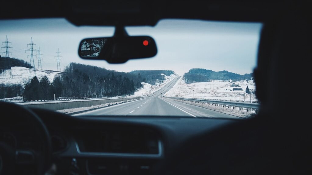 Vue depuis l’intérieur d’une voiture en hiver : tableau de bord sombre, rétroviseur, et une autoroute bordée de collines enneigées et de pylônes électriques