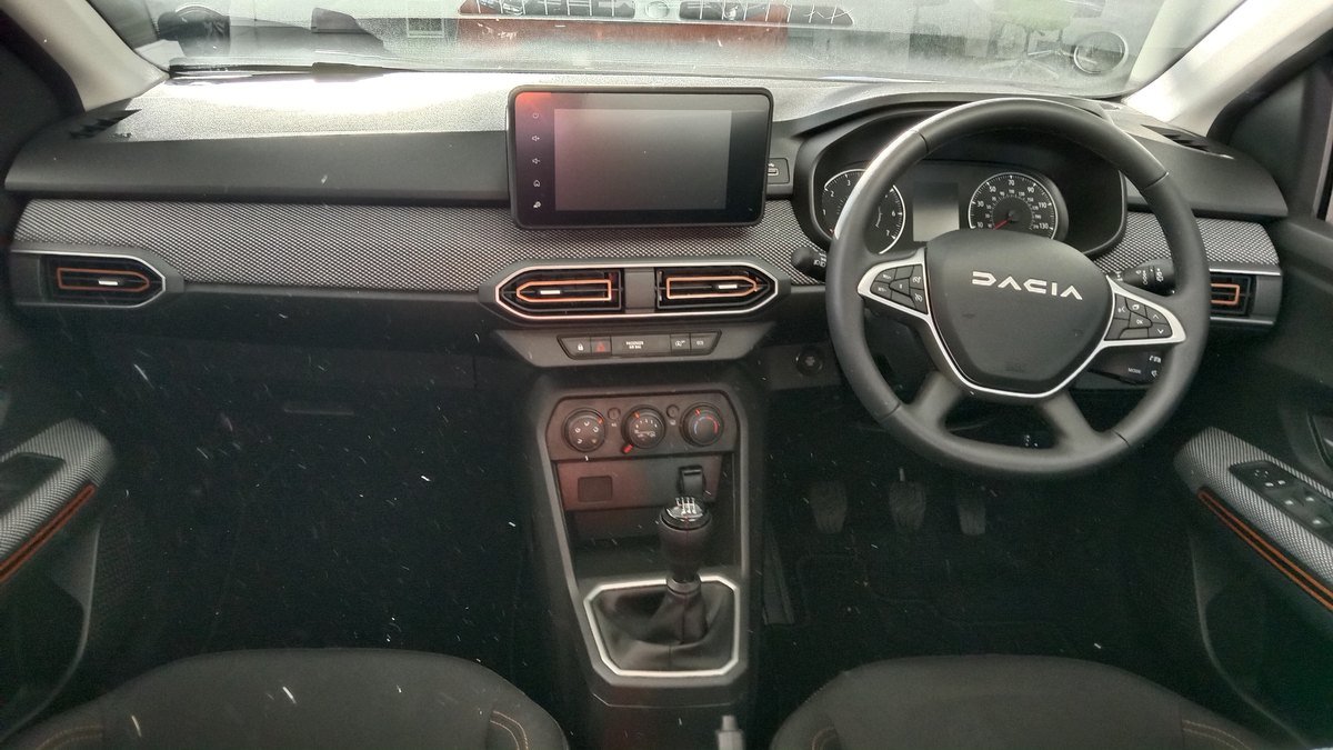 Tableau de bord d’une Dacia Sandero : petit écran tactile en haut, trois molettes rondes de climatisation sous les aérateurs, levier de vitesses manuel