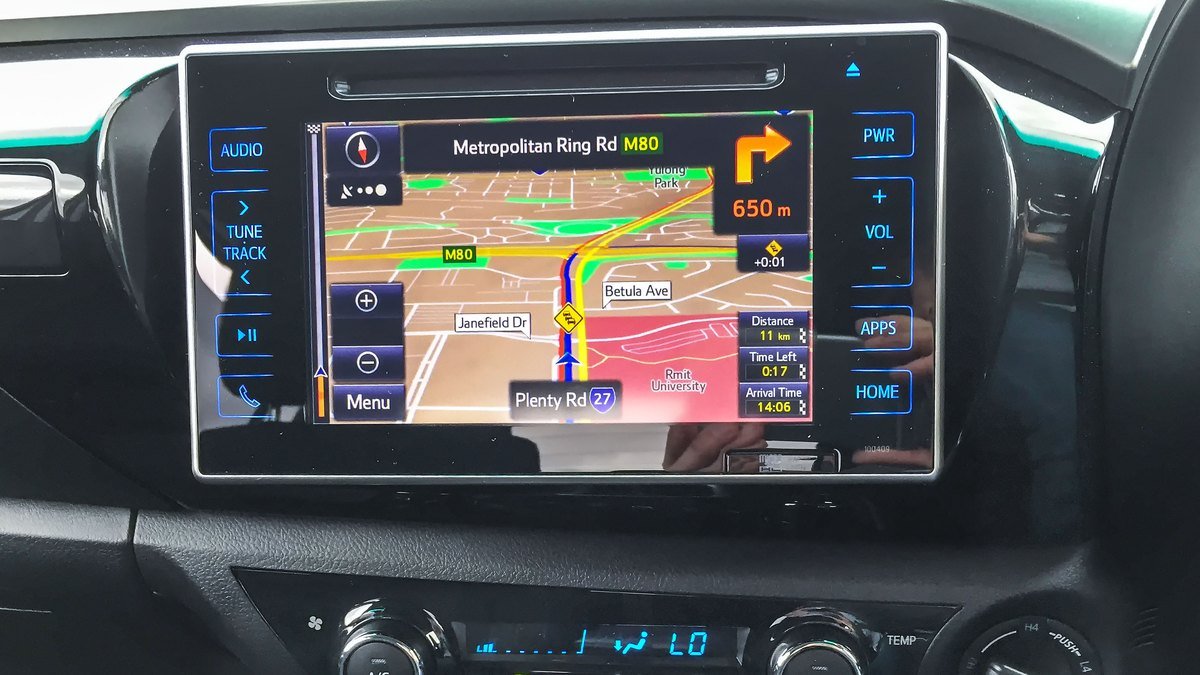 Écran tactile central d’un Toyota Hilux affichant une carte de navigation avec un itinéraire, des boutons de volume et de menu de chaque côté, et les commandes de climatisation en dessous