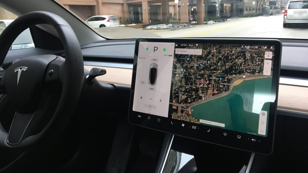 Tableau de bord presque sans bouton d’une Tesla Model 3 : grand écran tactile central affichant une carte, volant à gauche, rue en arrière-plan