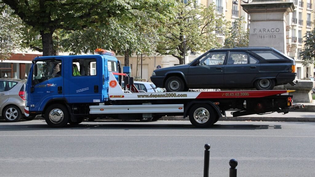 Camion-plateau bleu de dépannage transportant une voiture sombre dans une rue de Paris bordée d’arbres et d’immeubles haussmanniens