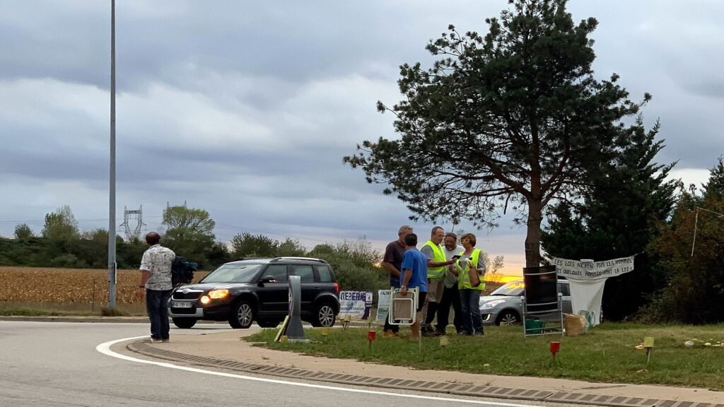 Quelques personnes en gilet jaune réunies au bord d’un rond-point à la sortie d’autoroute de Meximieux-Pérouges, pendant qu’un SUV noir passe phares allumés sous un ciel gris