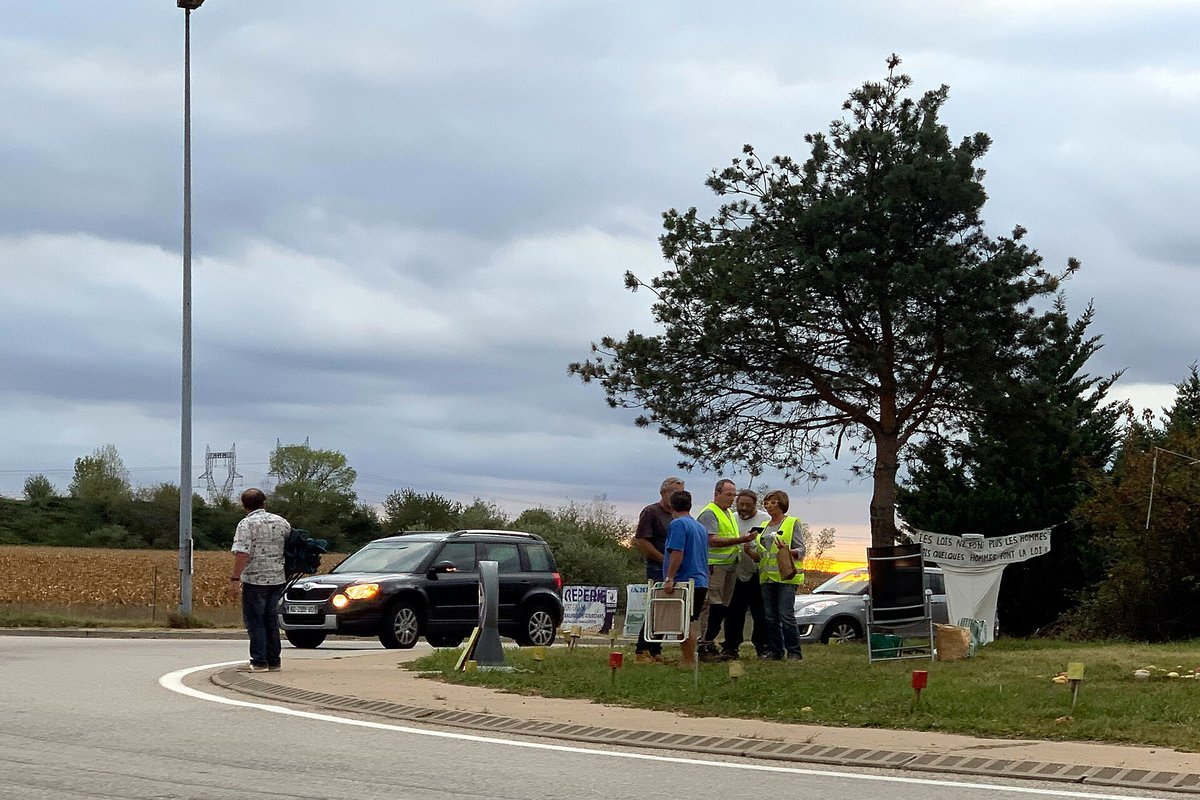 Quelques personnes en gilet jaune rassemblées sur un rond-point en sortie d’autoroute, avec des banderoles accrochées à une barrière et une voiture qui passe
