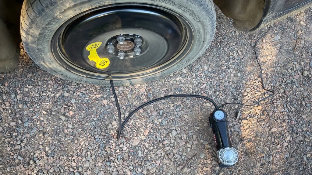 Petit compresseur 12 V noir posé sur le gravier, son tuyau vissé sur la valve d’une roue de secours galette montée sur une voiture