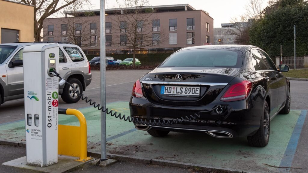 Mercedes-Benz noire de type hybride rechargeable garée sur une place verte et reliée par un câble spiralé à une borne de recharge blanche, devant un immeuble