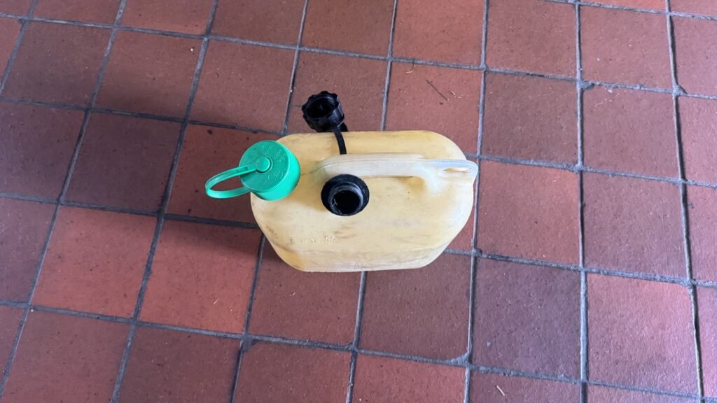 Jerrican plastique jaune de 5 litres (mention max. 5 L) posé sur un carrelage de garage, bouchon noir détaché — illustration