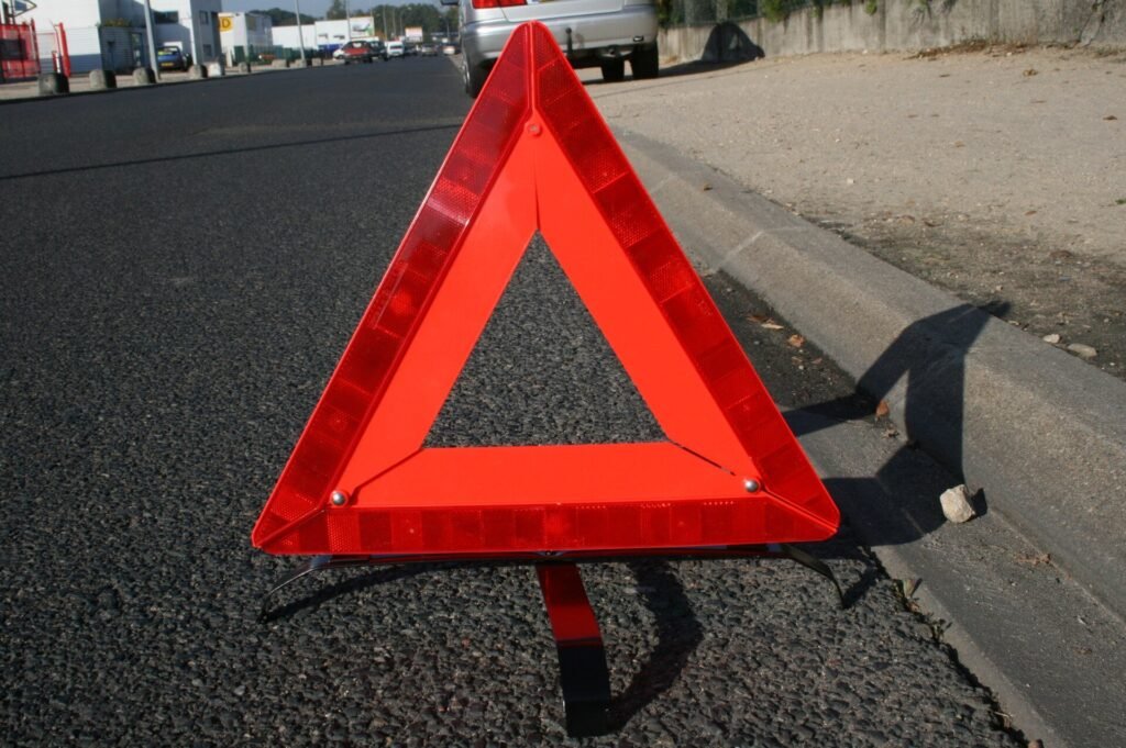Triangle de présignalisation rouge et réfléchissant posé sur l’asphalte, au bord d’une route, avec une voiture au loin