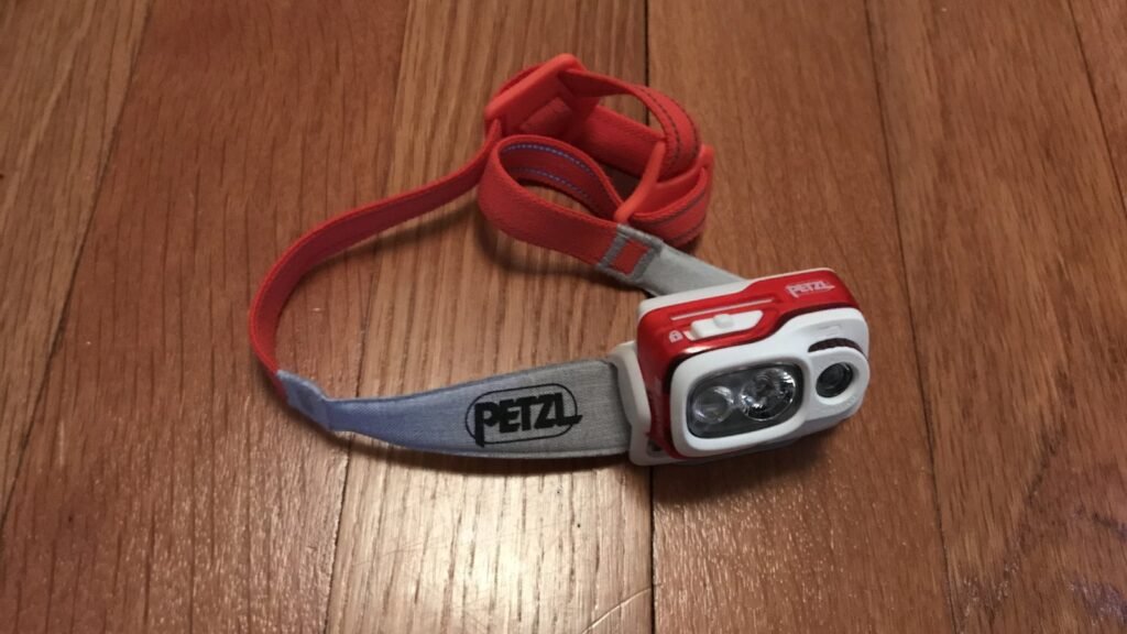 Lampe frontale Petzl rouge et grise, bandeau élastique déplié, posée sur un parquet en bois