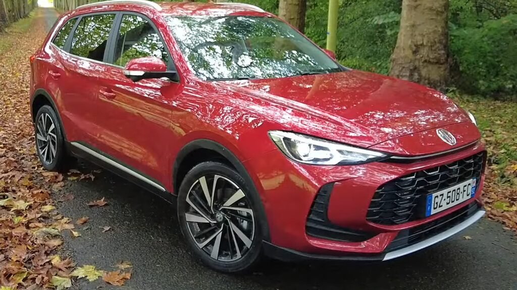 Un SUV MG ZS Hybrid+ rouge, plaque française, sur une route d’automne bordée d’arbres et de feuilles mortes