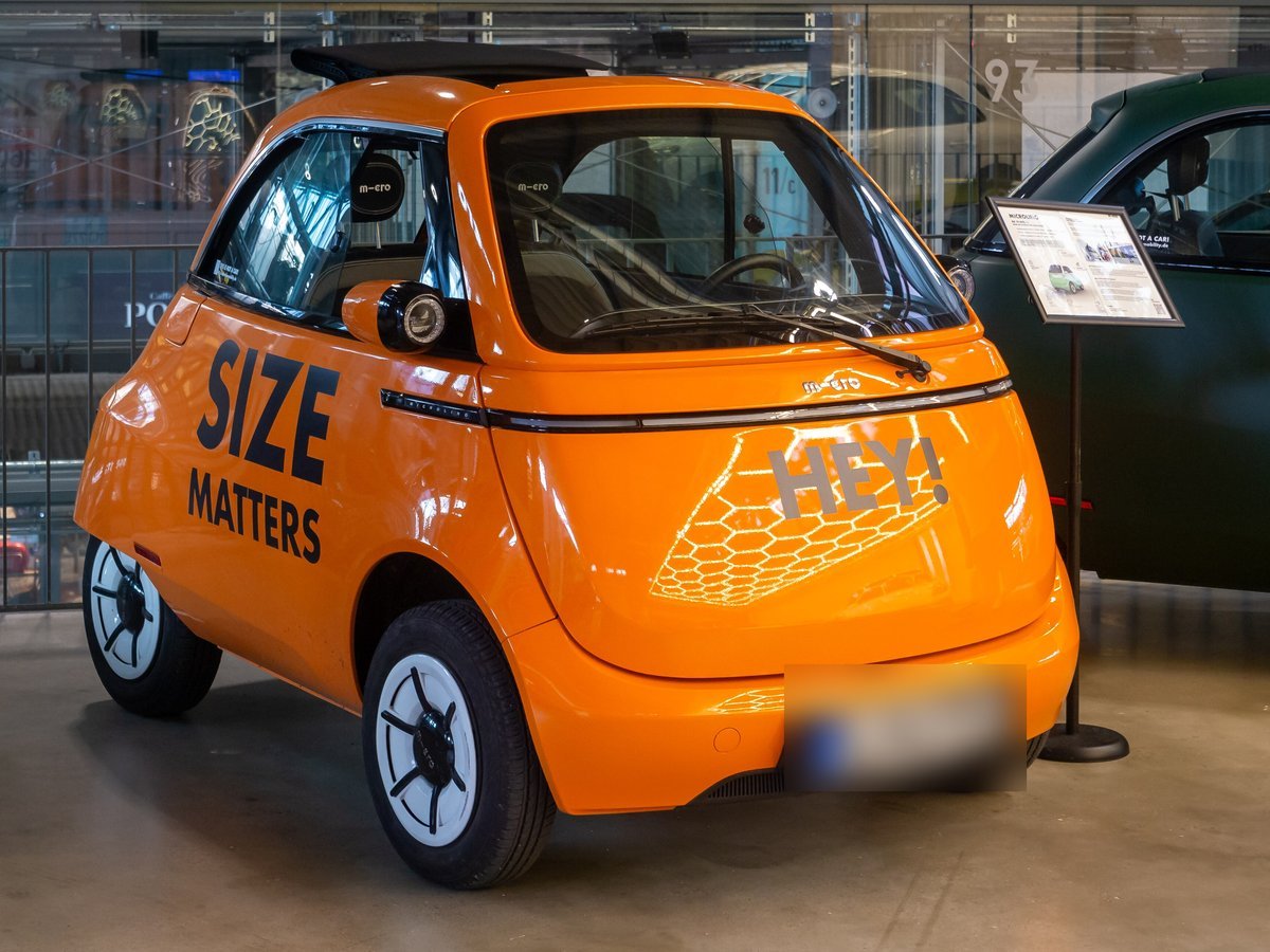 Microlino orange à portière frontale, petite voiture électrique en forme de bulle, exposée dans un hall avec un panneau d’information à côté