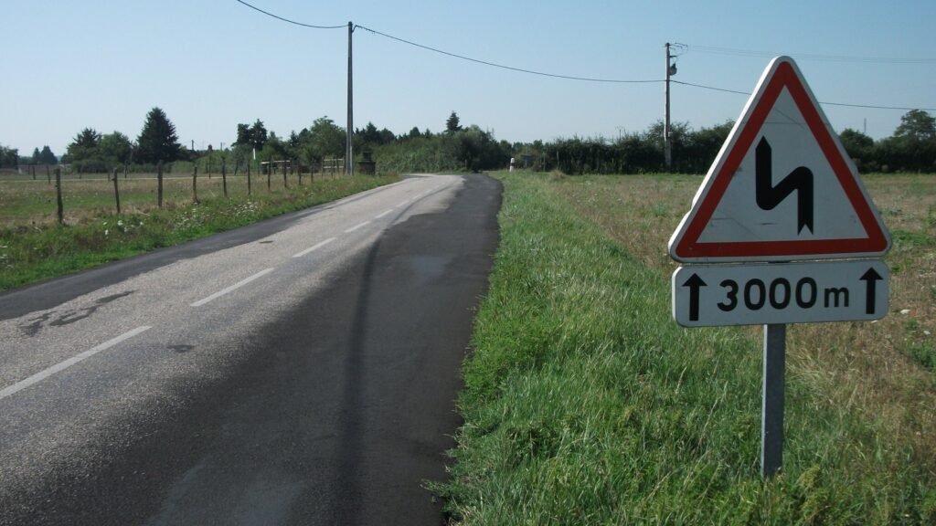Route départementale de campagne bordée de prés et de poteaux électriques, avec au premier plan un panneau triangulaire rouge annonçant une succession de virages sur 3 000 mètres, sous un ciel bleu