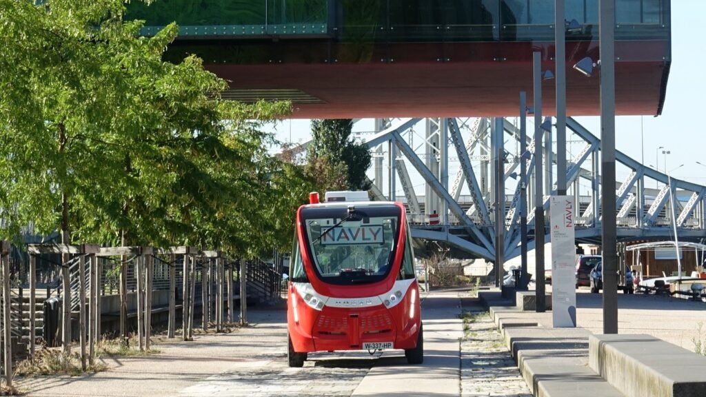 Navette autonome électrique rouge Navly roulant sur un quai piéton de Lyon Confluence, sous un immeuble vitré et devant un pont métallique