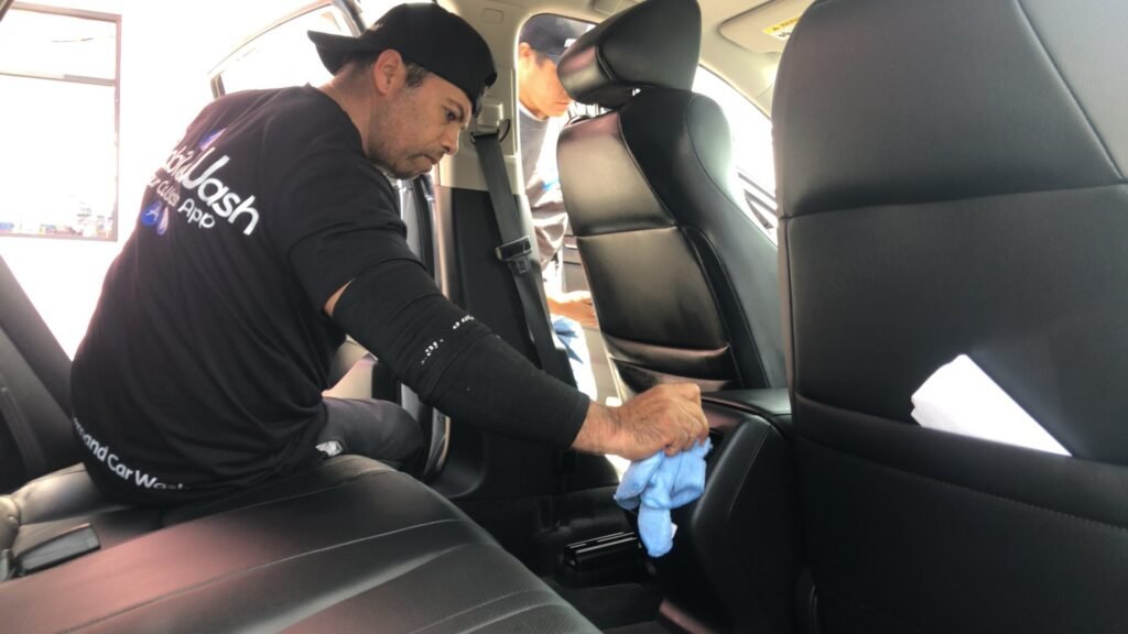 Un professionnel essuie avec un chiffon microfibre bleu le dossier d’un siège en cuir noir, à l’intérieur d’une voiture portières ouvertes