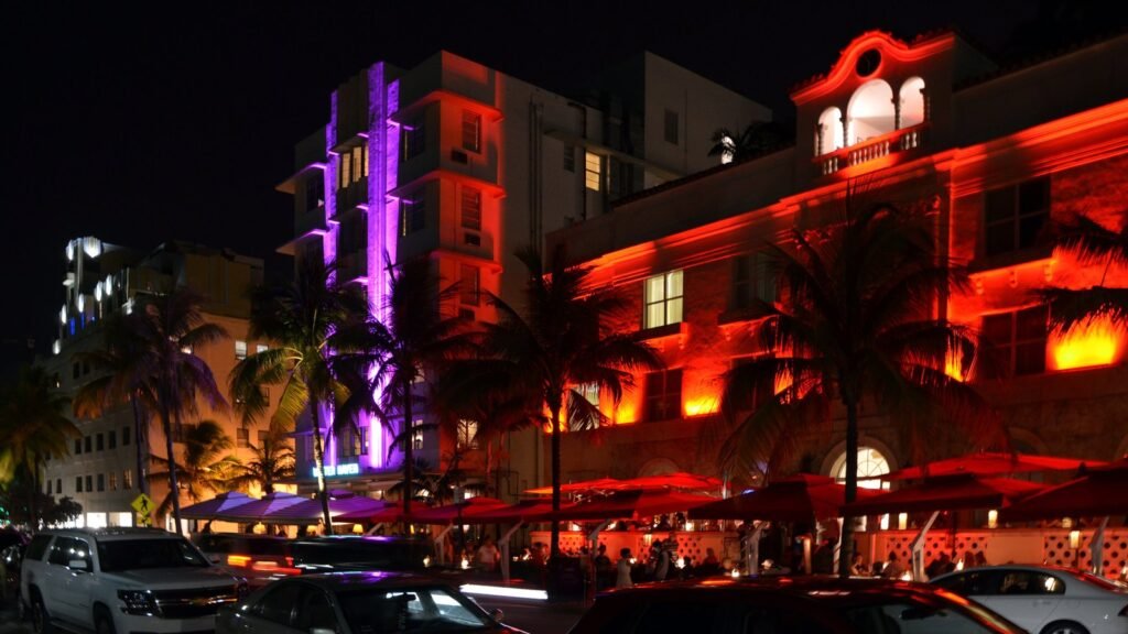 Façades Art déco d’Ocean Drive à Miami Beach illuminées la nuit de néons violets et orange, avec des palmiers, des terrasses et des voitures garées au premier plan
