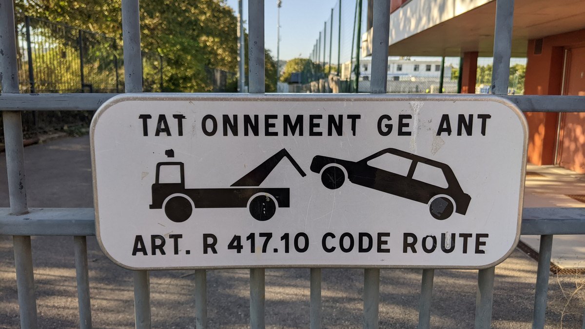 Panonceau blanc « stationnement gênant » fixé à une grille à Lyon, avec le dessin d’une dépanneuse qui enlève une voiture et la mention « Art. R 417.10 Code route » ; des lettres arrachées le font lire « tatonnement geant »