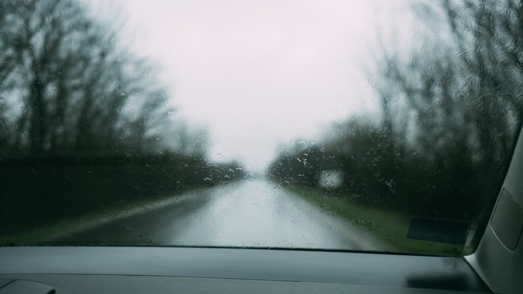 Vue depuis le siège conducteur : pare-brise couvert de gouttes et de buée, route de campagne bordée d’arbres sans feuilles sous un ciel gris, tableau de bord au premier plan