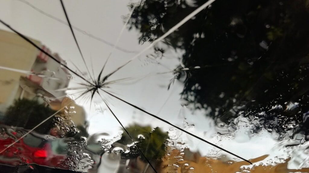 Pare-brise fissuré vu de l’intérieur de la voiture : impact en étoile d’où partent de longues fissures, gouttes de pluie et arbres en arrière-plan