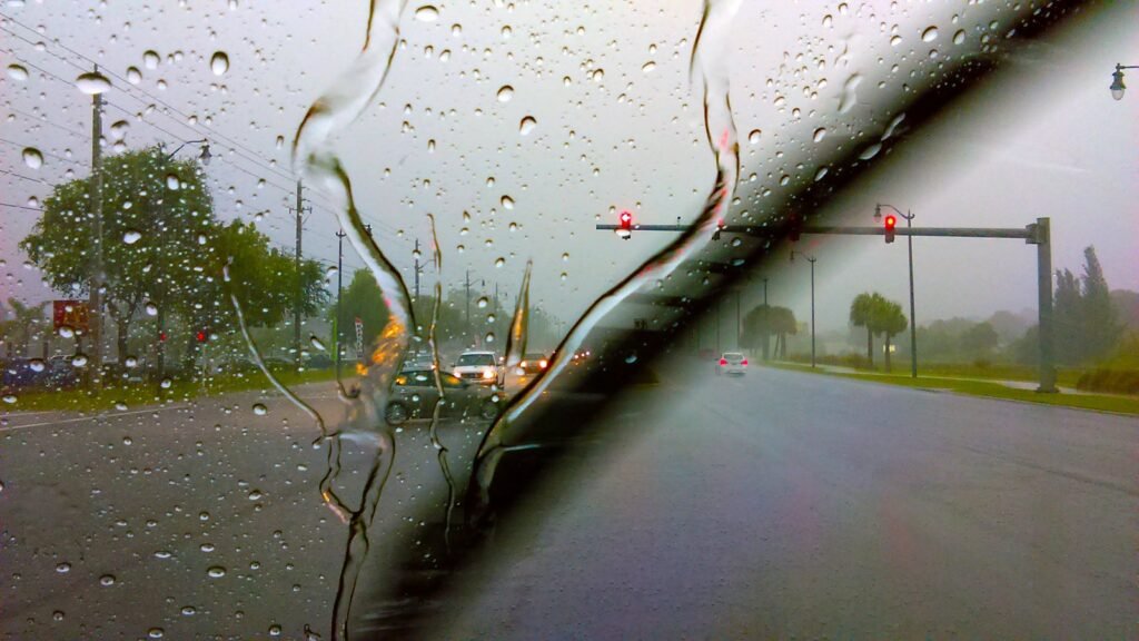 Vue depuis l’habitacle à travers un pare-brise couvert de gouttes de pluie, avec un balai d’essuie-glace en mouvement et un feu rouge au loin sur une route urbaine