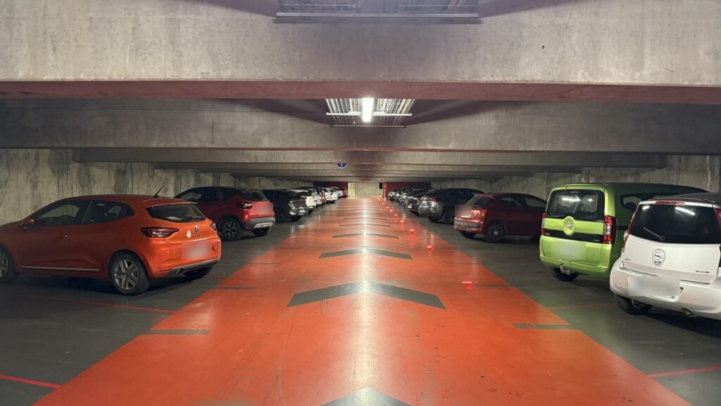 Allée d’un parking souterrain aux voies orange, avec des voitures stationnées de chaque côté sous un plafond en béton éclairé