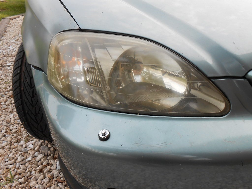 Gros plan sur un phare avant de Honda Civic grise : la glace en plastique est jaunie et voilée, on distingue mal le réflecteur
