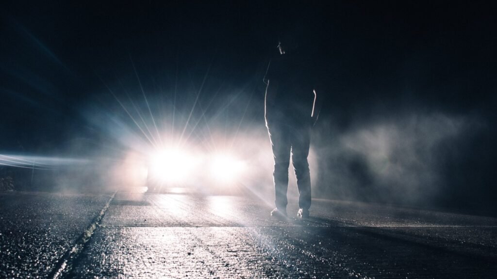 Deux phares de voiture très lumineux éblouissent la nuit sur une route mouillée, face à une silhouette debout dans la brume