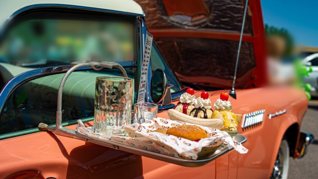 Plateau de drive-in rétro accroché à la vitre d’une voiture de collection orange, avec burger, banana split et verres, sous un grand soleil