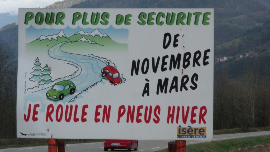 Panneau routier en Isère : « Pour plus de sécurité, de novembre à mars, je roule en pneus hiver », avec deux voitures dessinées sur une route de montagne