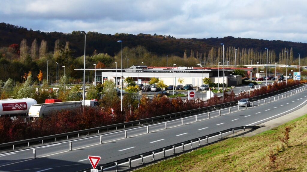 Autoroute A89 en Dordogne bordée d’arbustes rouges d’automne, avec une voiture et un camion-citerne, et en arrière-plan l’aire de services du Manoire avec son parking et ses collines boisées
