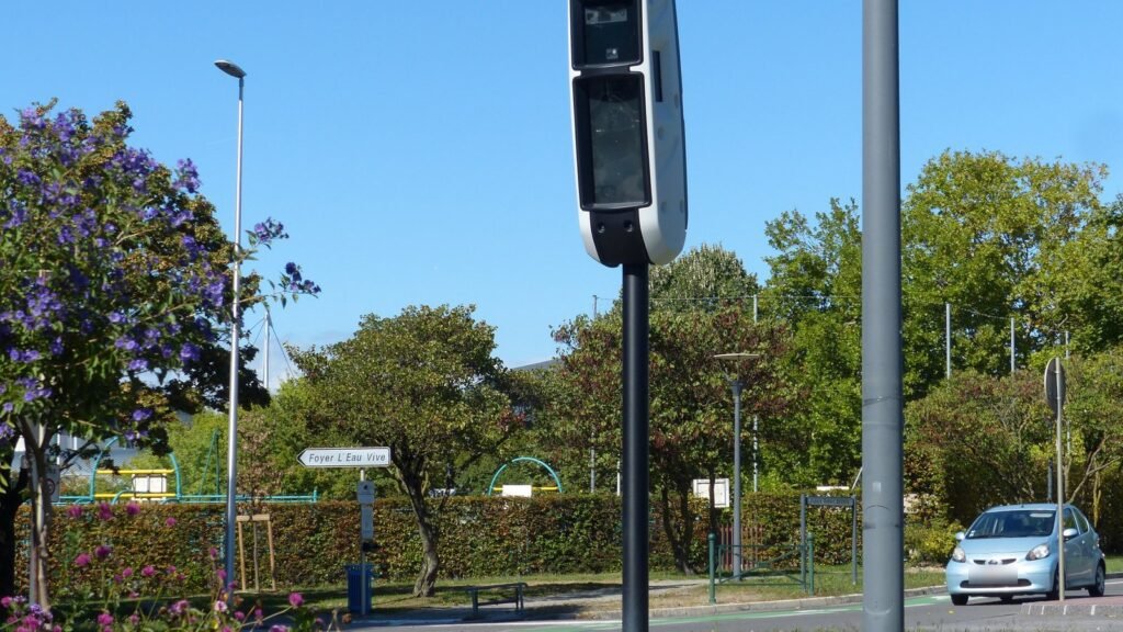 Radar tourelle noir monté sur un mât au bord d’une avenue d’Annemasse, avec des arbres, un massif fleuri et une voiture qui passe en arrière-plan