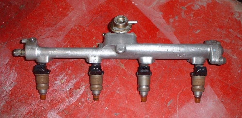 Rampe d’injection d’essence en métal avec quatre injecteurs et un régulateur de pression, posée sur un fond rouge