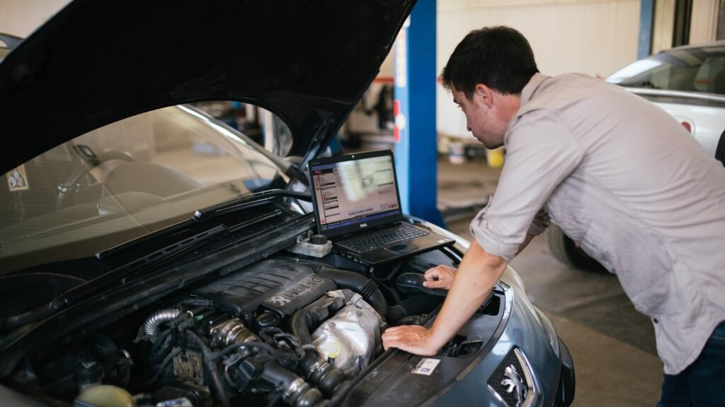 Mécanicien penché sous le capot ouvert d’une Peugeot grise, ordinateur portable de diagnostic posé sur le moteur, dans un atelier