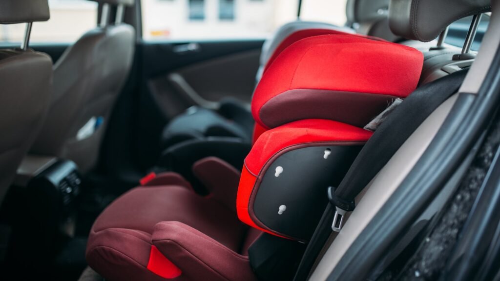 Rehausseur enfant avec dossier rouge et bordeaux, appuie-tête et protections latérales, installé sur la banquette arrière d’une voiture, à côté de la ceinture de sécurité