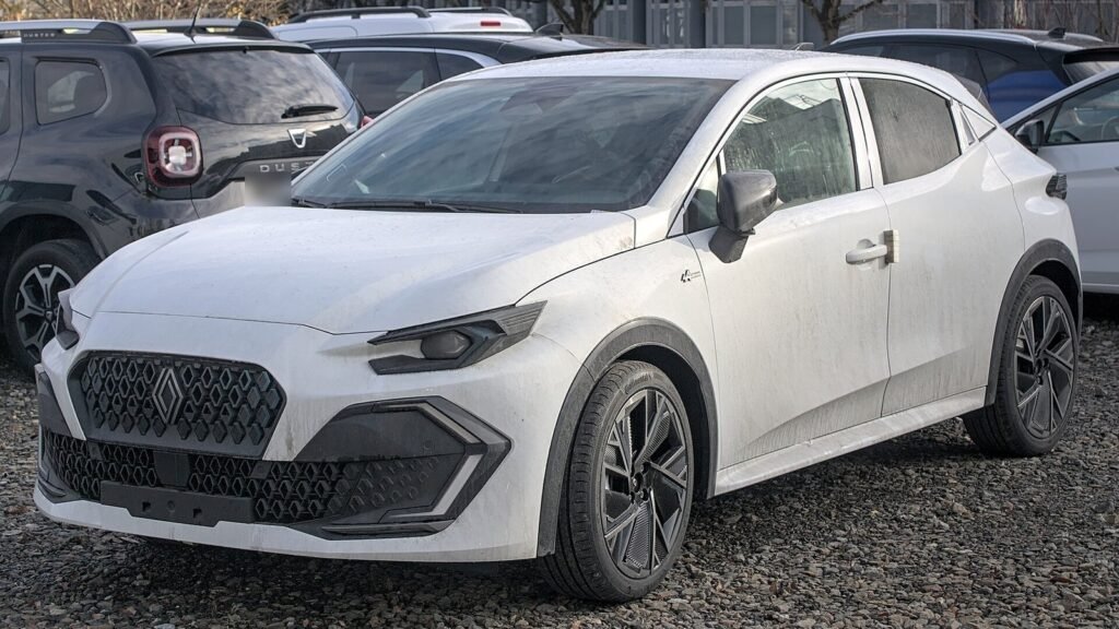 Renault Clio de sixième génération blanche, vue de trois quarts avant, garée sur un parking en gravier entre d’autres voitures, dont un Dacia Duster gris foncé à la plaque floutée