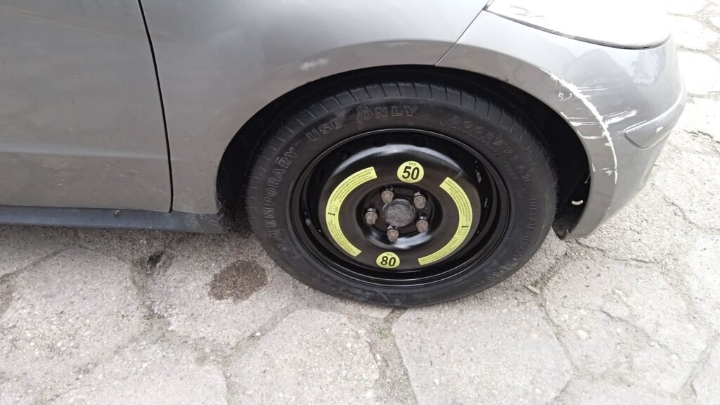 Roue de secours galette à jante noire montée à l’avant d’une voiture grise, avec la mention Temporary use only et des étiquettes jaunes de vitesse maximale (50 et 80)