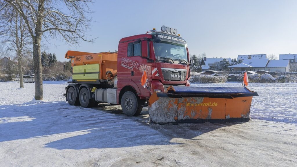 Camion de déneigement équipé d’une lame orange et d’une saleuse sur une place enneigée, illustration du salage des routes en hiver