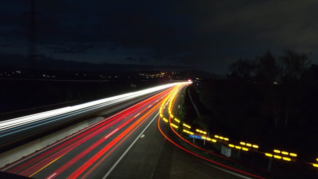 Autoroute la nuit en pose longue : traînées blanches des phares et rouges des feux arrière dans un virage, balises jaunes sur le bas-côté