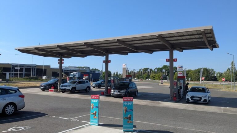 Station-service TotalEnergies sous un grand auvent, plusieurs voitures au plein, totem de prix et ciel bleu — aire autoroutière en France